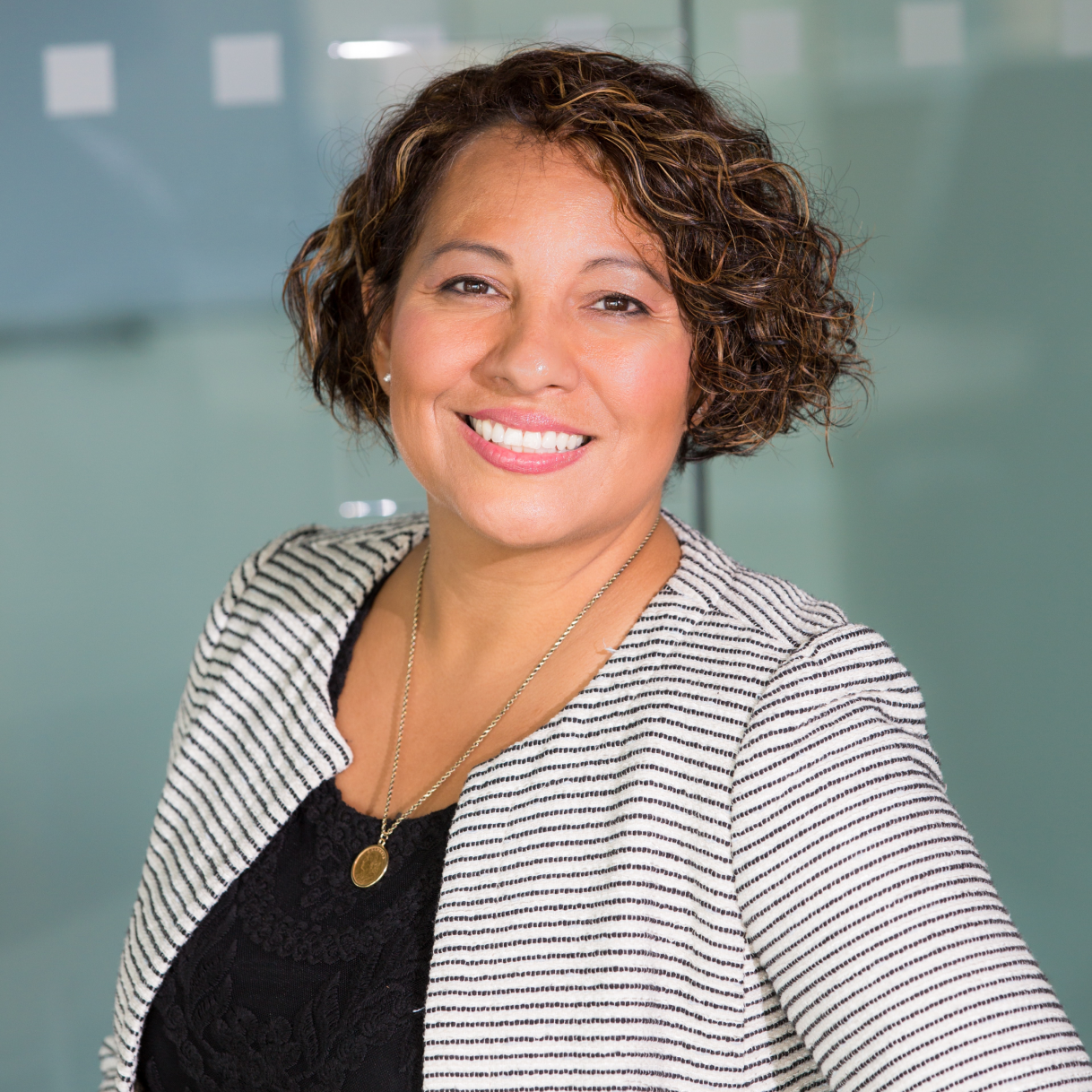 A smiling woman with curly hair, wearing a striped blazer and necklace, poses in an office setting with a glass background.