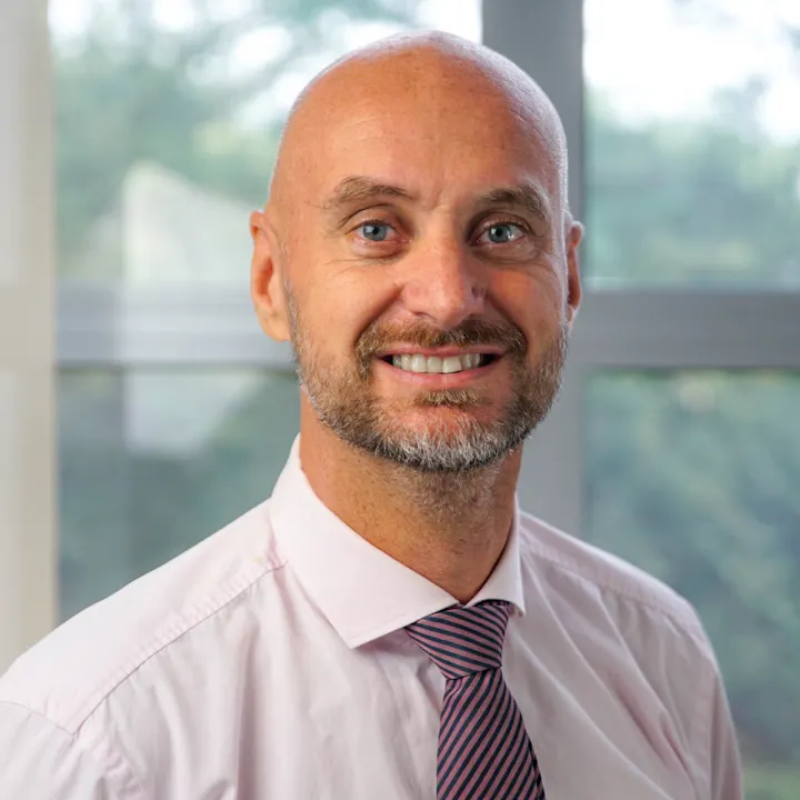 A smiling man with a shaved head, beard, and blue eyes, wearing a pink shirt and striped tie against a bright background.