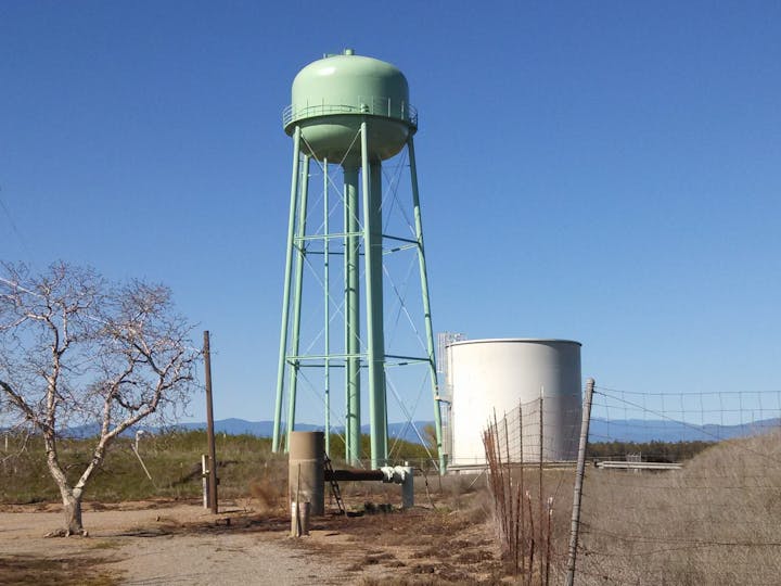 The image features a green water tower and a white storage tank against a clear blue sky, with sparse vegetation nearby.