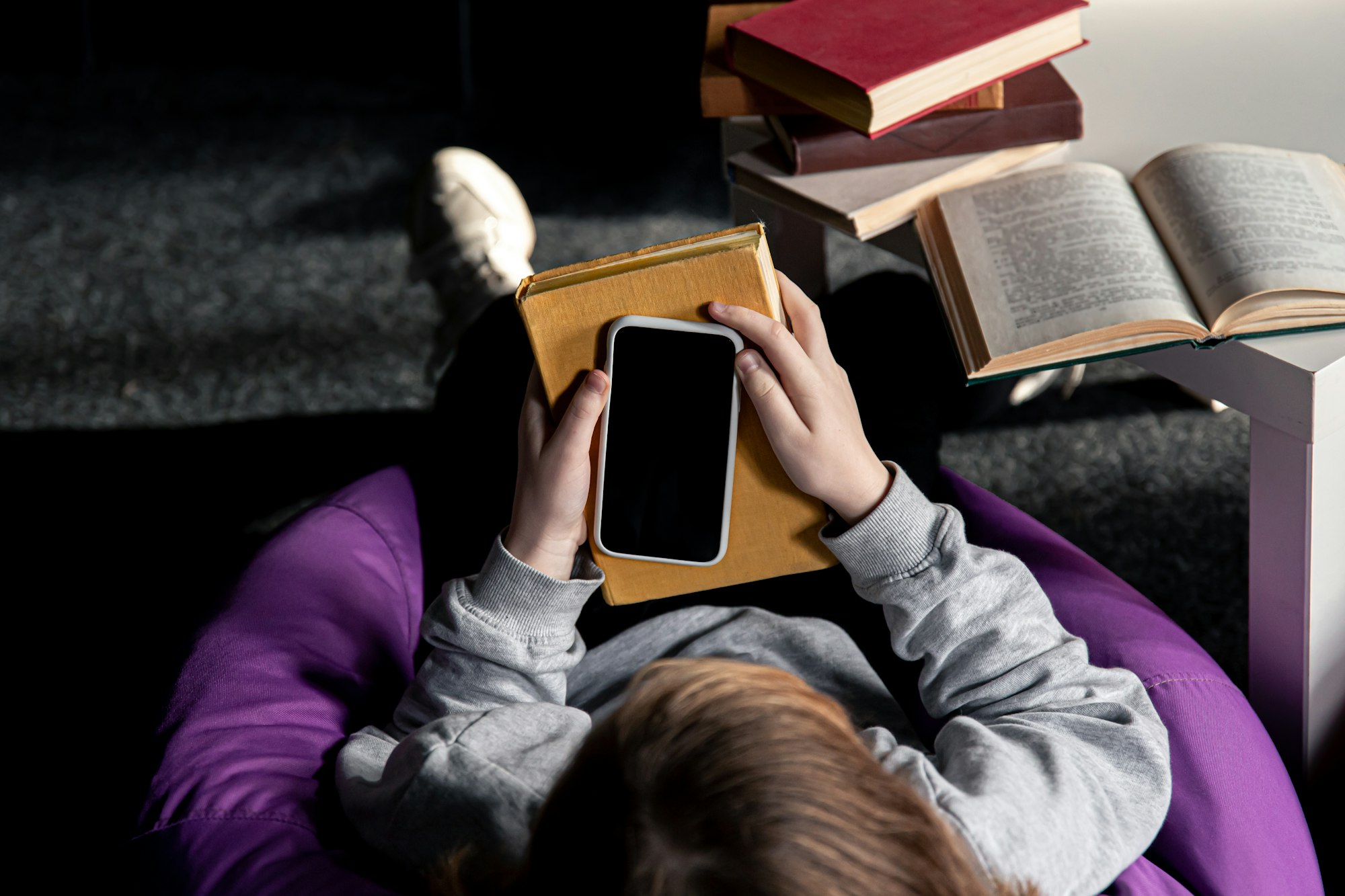 A person sits in a bean bag, holding a book and a smartphone, with stacked books and an open book nearby.