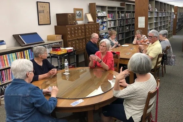 A group of people sitting around tables in a library, engaged in conversation or activities.