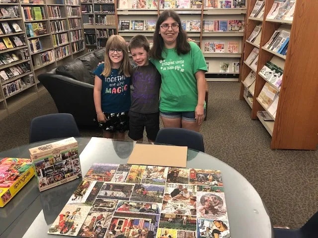 Three smiling people in a library with a completed puzzle on a table.