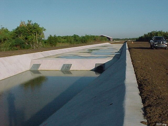 A concrete drainage canal filled with water runs alongside a dirt road with a parked truck, surrounded by greenery.