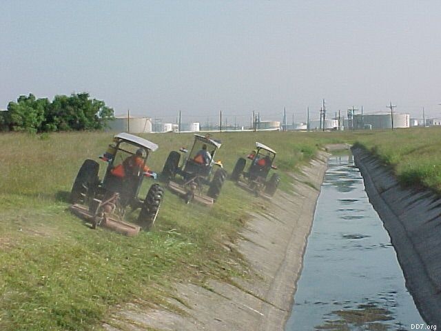 Tractors mowing grass on a sloped canal bank near an industrial area with storage tanks in the background.