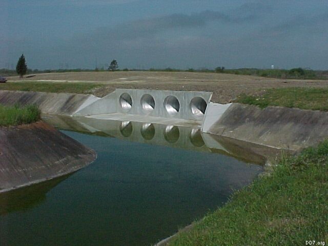 A concrete structure with four culverts beneath a grassy embankment, partially submerged in water.