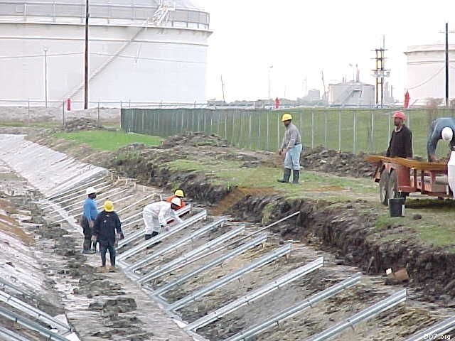 Workers in hard hats installing metal frames in a sloped trench near large storage tanks and a fence.