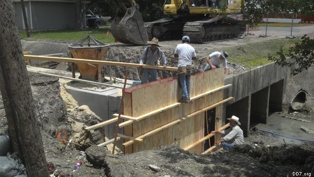 Construction site with workers building a wooden structure near a culvert, alongside heavy machinery and concrete forms.