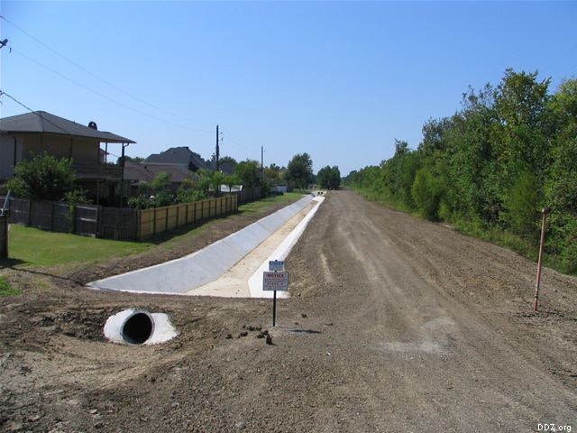 A dirt road with a concrete drainage ditch, housing on the left, trees on the right, and a sign next to the ditch outlet.