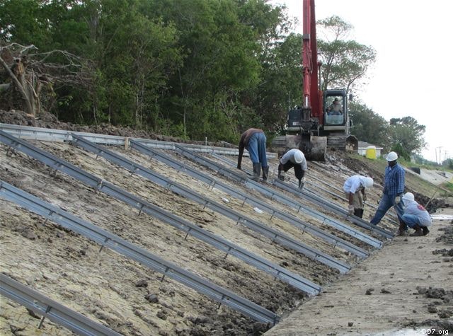 Workers in hard hats installing metal beams on a sloped embankment near construction machinery and trees.