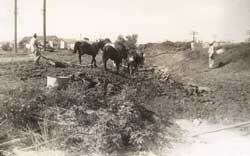 Two horses plowing a field, with people working around them; rural landscape setting.