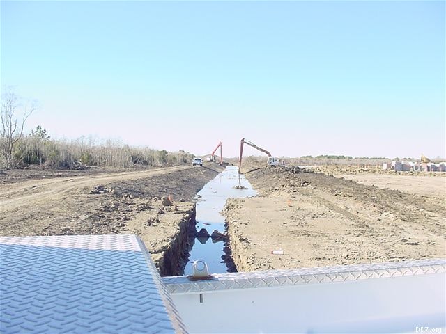 Construction site with a long trench filled with water, flanked by dirt piles and machinery in the distance.