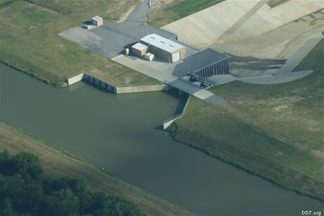 Aerial view of a water control structure with a building, gates, and a channel surrounded by grassy areas.