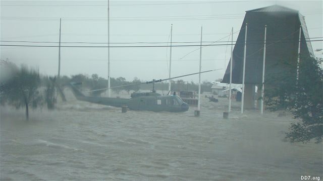Flooded area with a helicopter partially submerged, near a large building and poles.