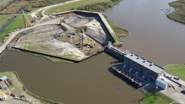 Aerial view of a construction site with heavy machinery near a body of water and a large building with industrial structures.