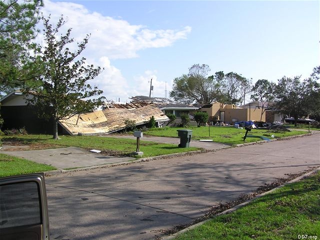 Street scene with house roofs collapsed, debris scattered, and multiple trees around.