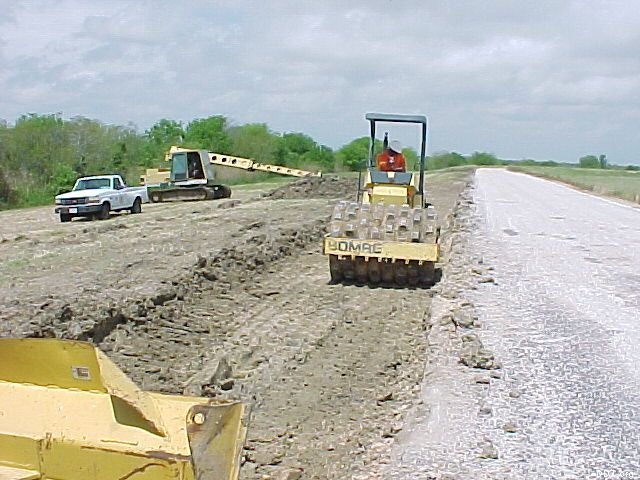 Construction site with heavy machinery, including a soil compactor and an excavator, working on a dirt road next to a pickup truck.
