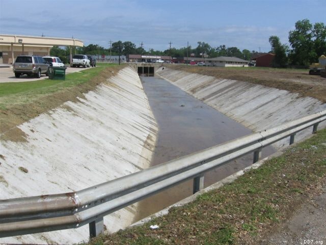 A drainage ditch with some water, surrounded by grass and a guardrail, located near a parking lot and buildings.