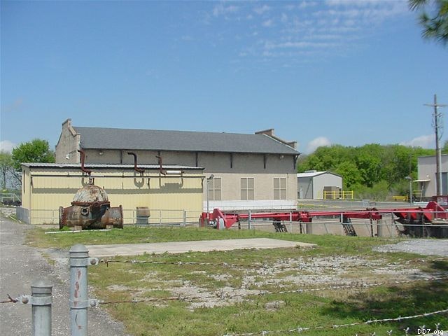 An industrial facility with a large building, storage tanks, pipes, and grassy area under a clear blue sky.