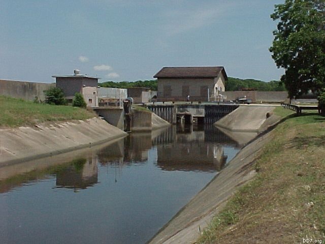 A canal with buildings and a dam structure, surrounded by grassy banks and trees under a clear sky.