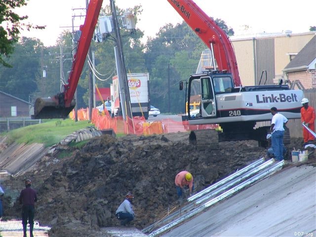 Construction site with workers and an excavator near a drainage area, alongside barriers and utility poles.