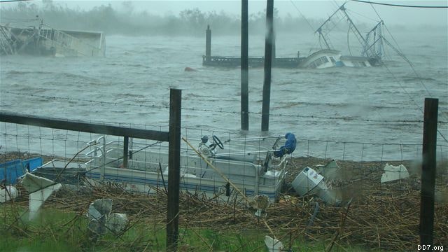 Flooded area with debris, including overturned boats and damaged fences, under gray, stormy skies.