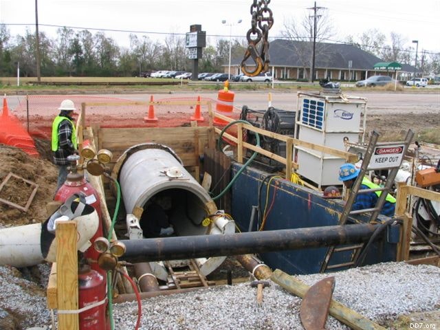 Construction site with workers, large pipe installation, safety cones, and equipment. Signs indicate restricted area.