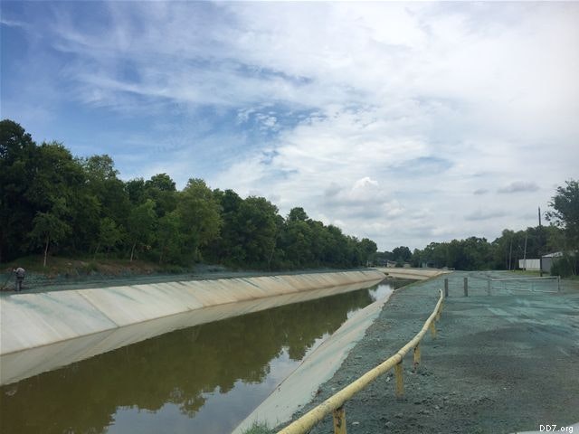 A concrete-lined canal with murky water runs alongside green grass and trees under a partly cloudy sky.