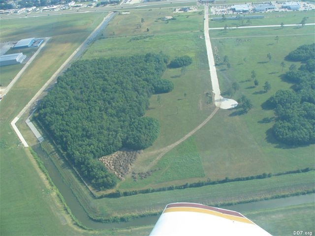 Aerial view of a landscape with fields, trees, roads, and a river or canal. Airplane wing visible in the foreground.