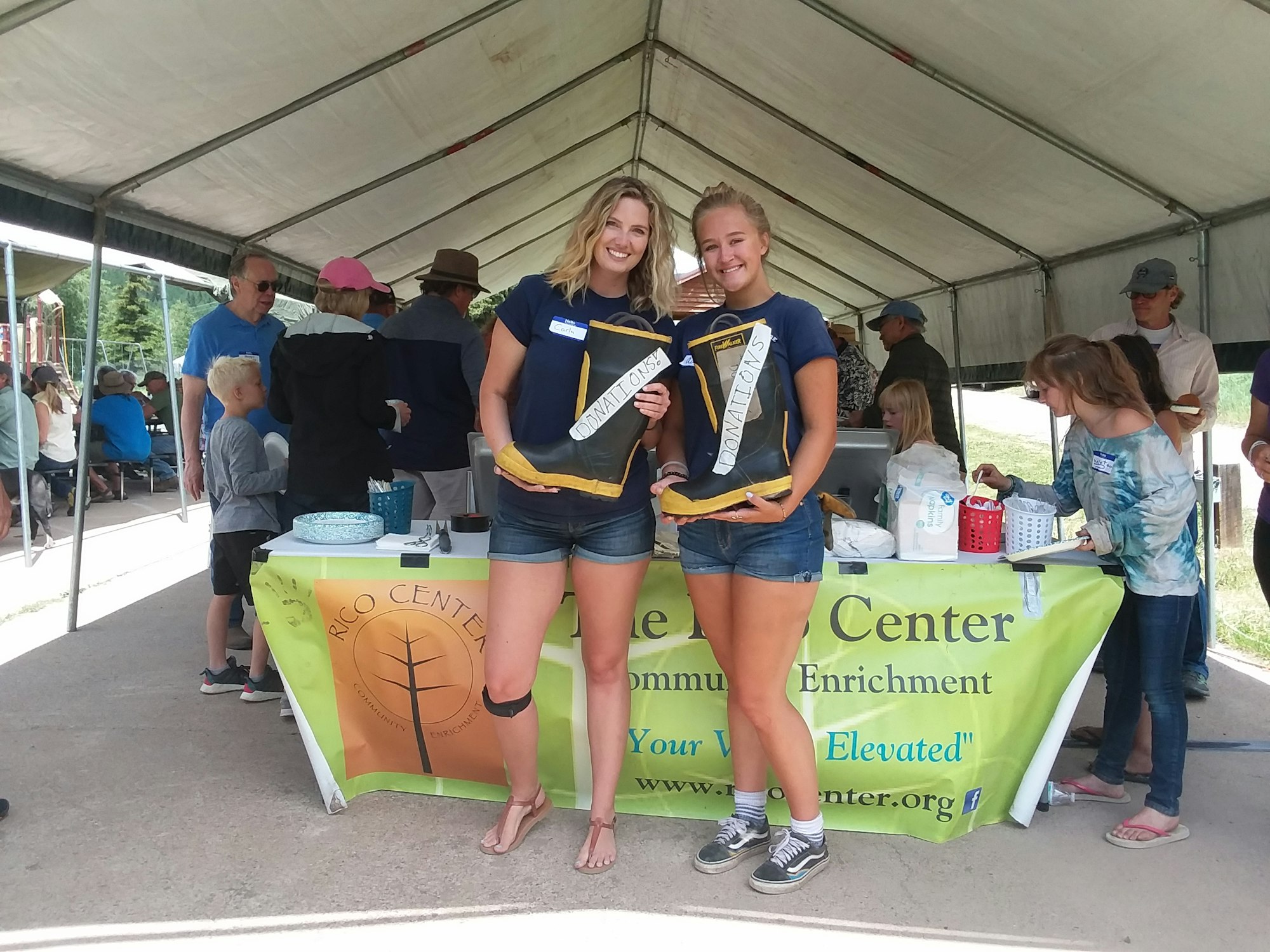 Two smiling people holding a sign, a table with a 'donations' jar, and other people in a tented area.
