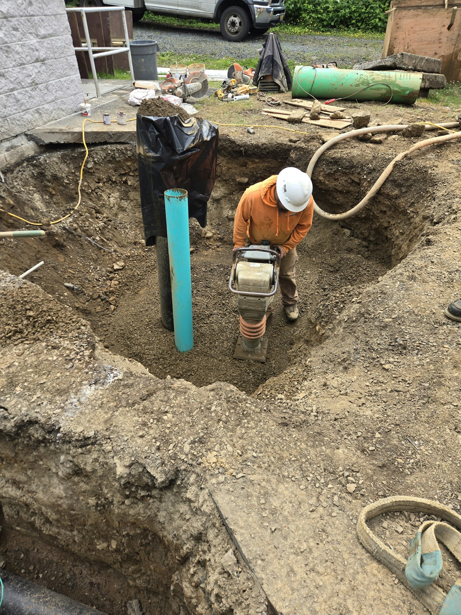 Construction worker with a hard hat operating equipment beside a blue pipe in a dirt excavation site.
