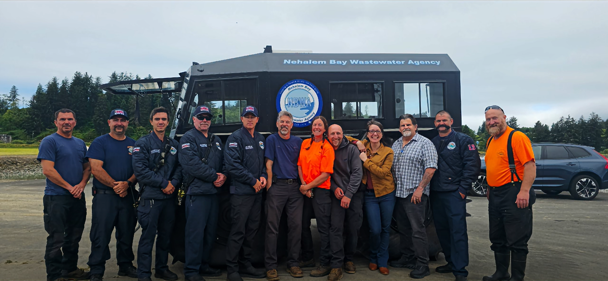 A group of people poses in front of a Nehalem Bay Wastewater Agency vehicle, showcasing teamwork in a scenic environment.