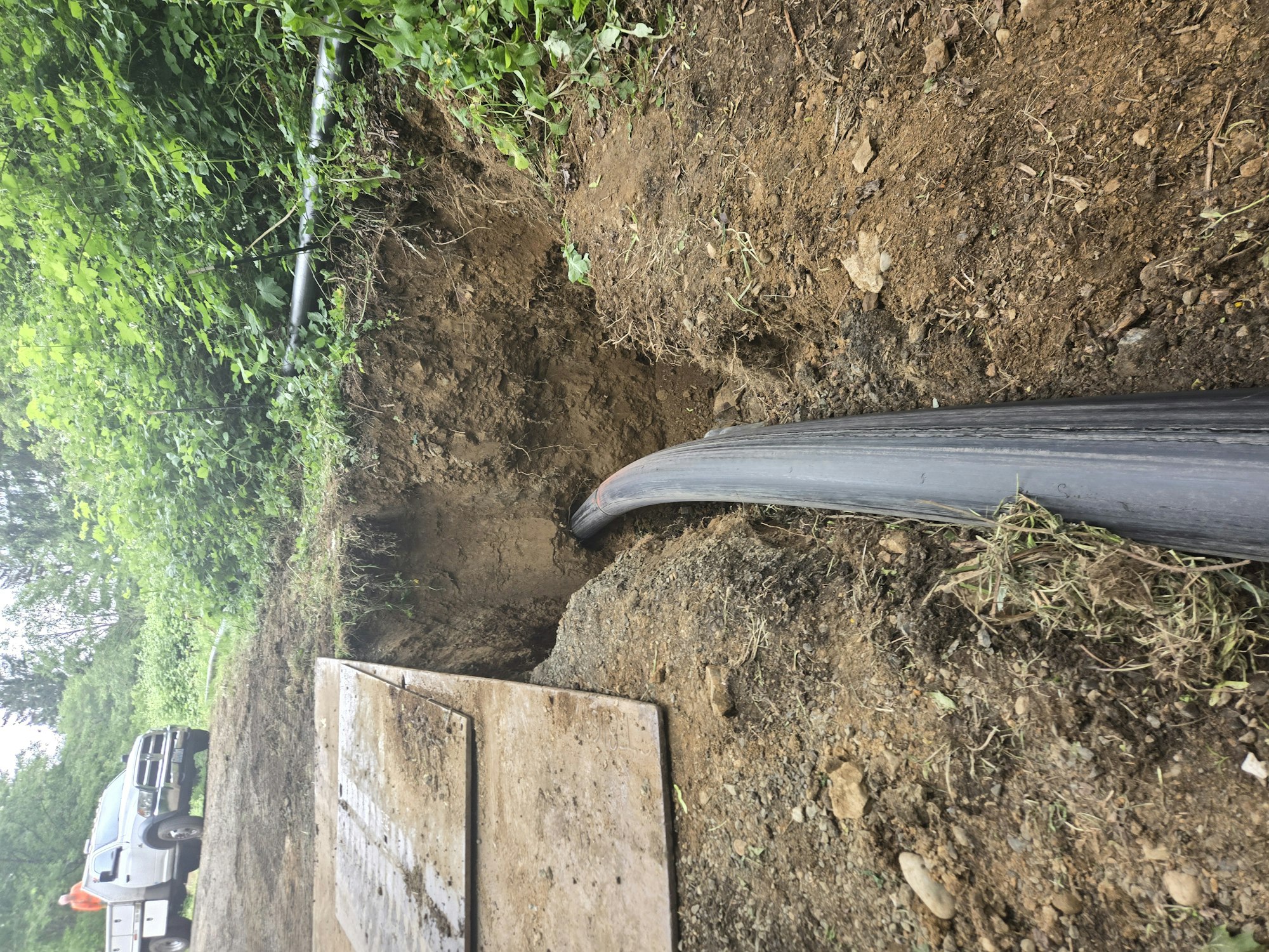 A pipe runs through a trench in the ground, with a truck visible in the background amidst greenery.