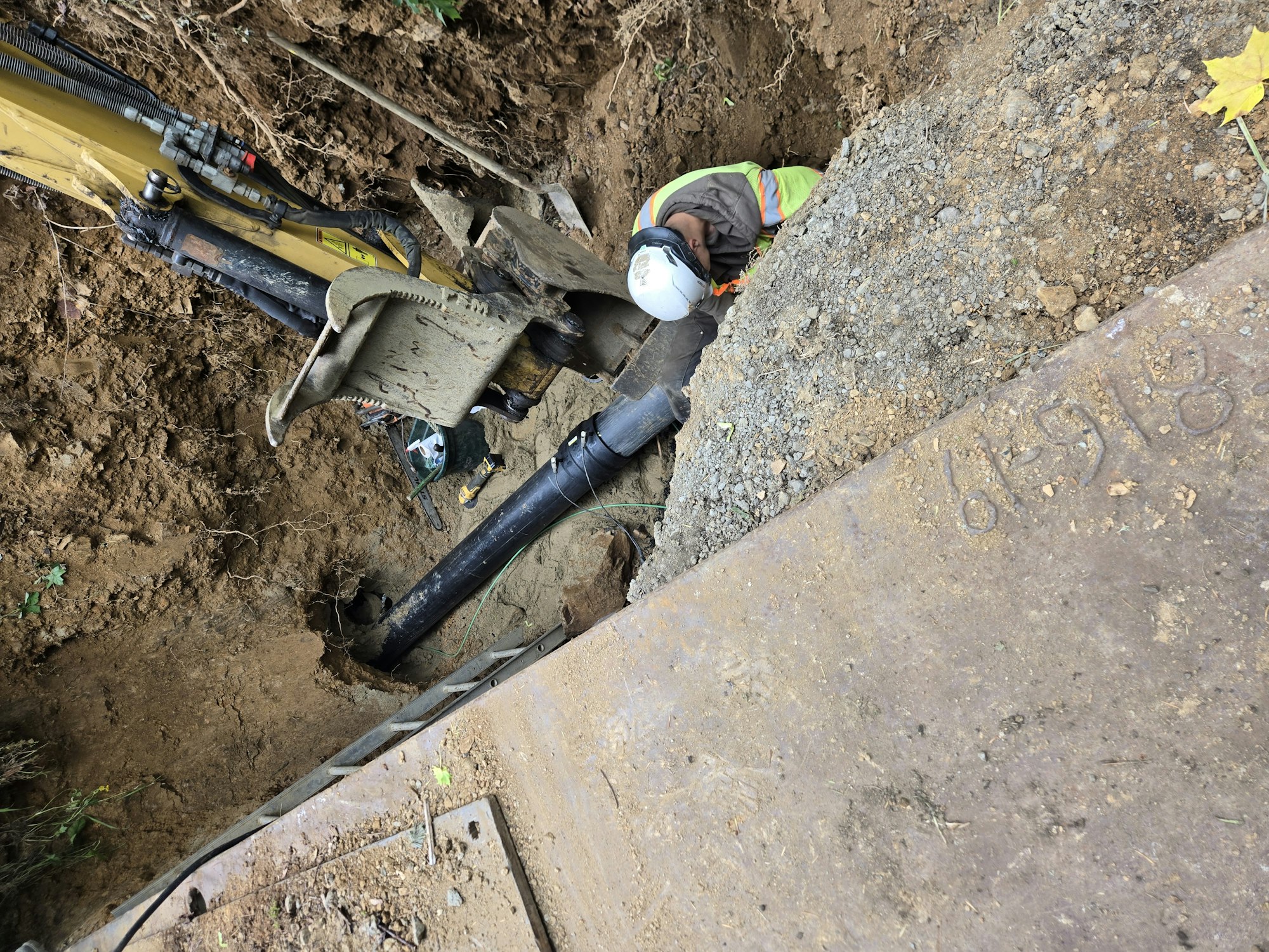 Construction worker in a trench, operating machinery near a black pipe, surrounded by dirt and excavated earth.