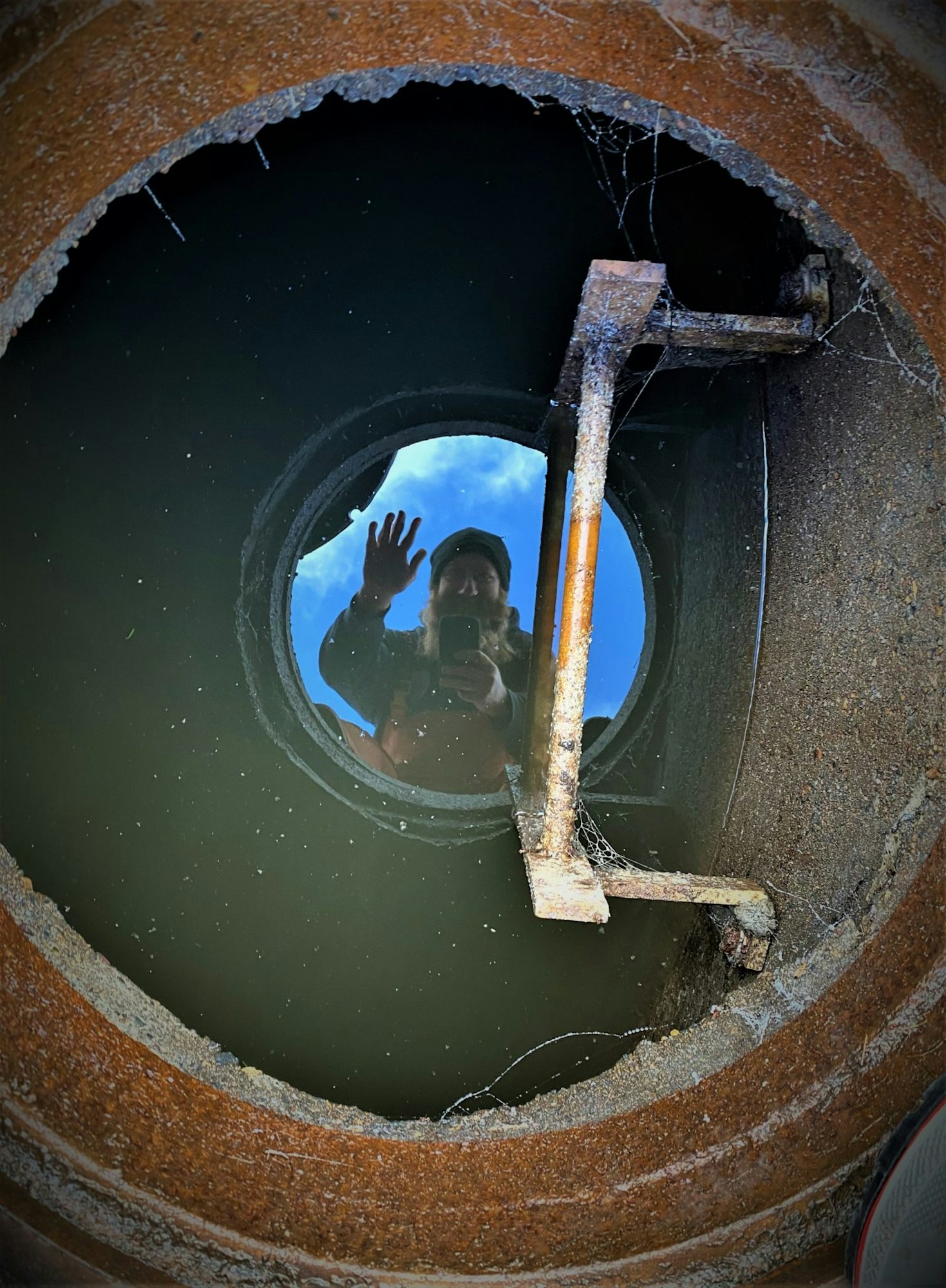 A person's reflection in water inside a circular rusty structure, with a hand raised in a wave, and a metal bar across.