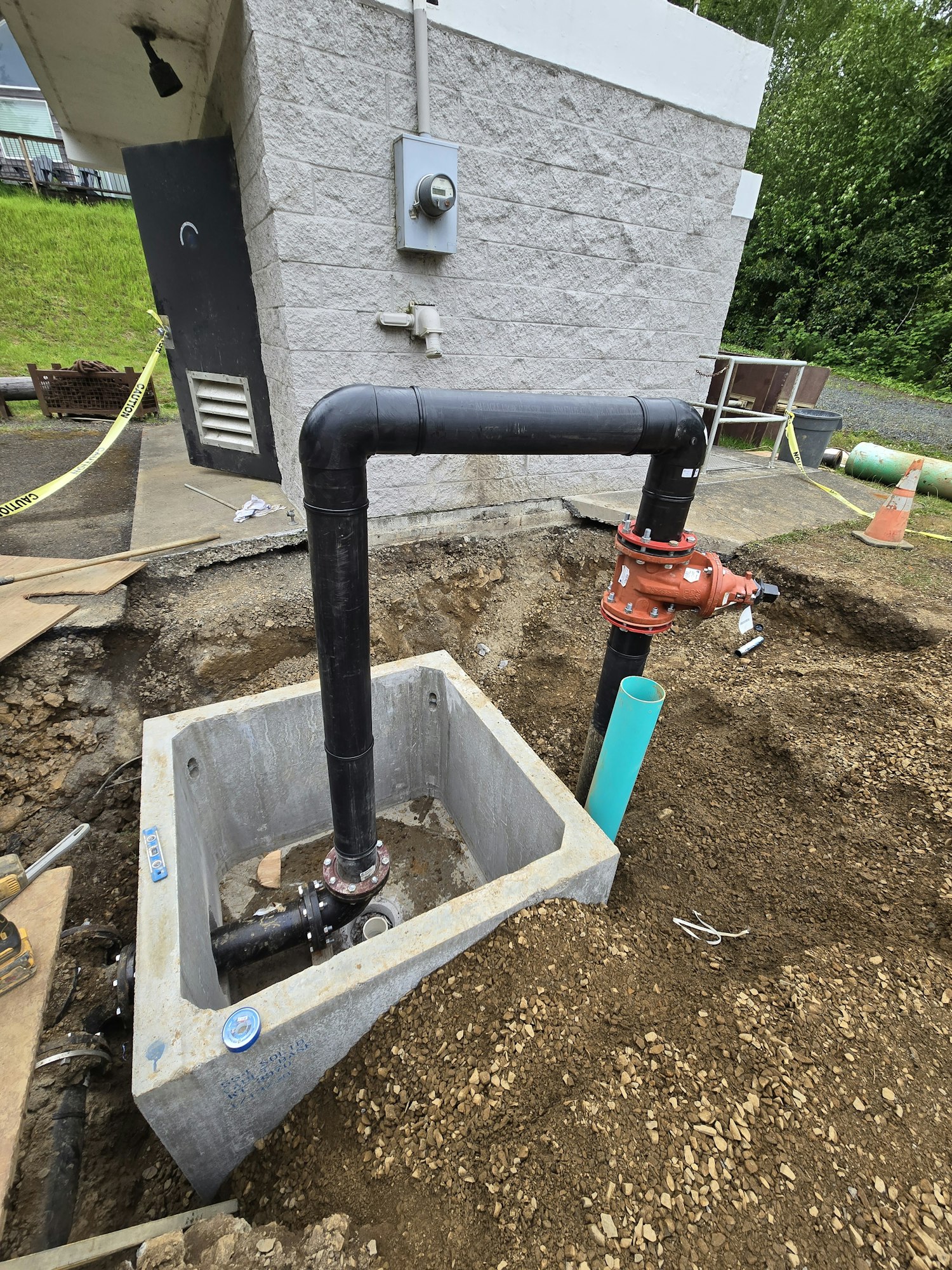 Construction site with pipes, a concrete box, gravel, and caution tape near a building wall.