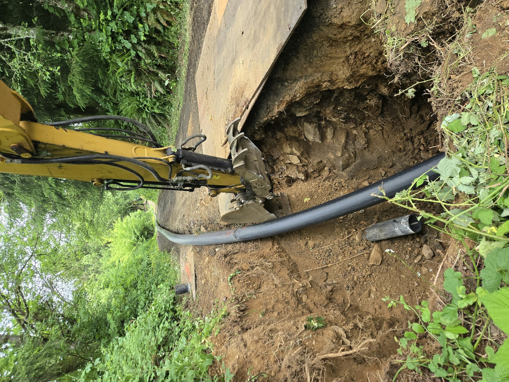 Excavator digging a trench for a black pipe in a forested area.
