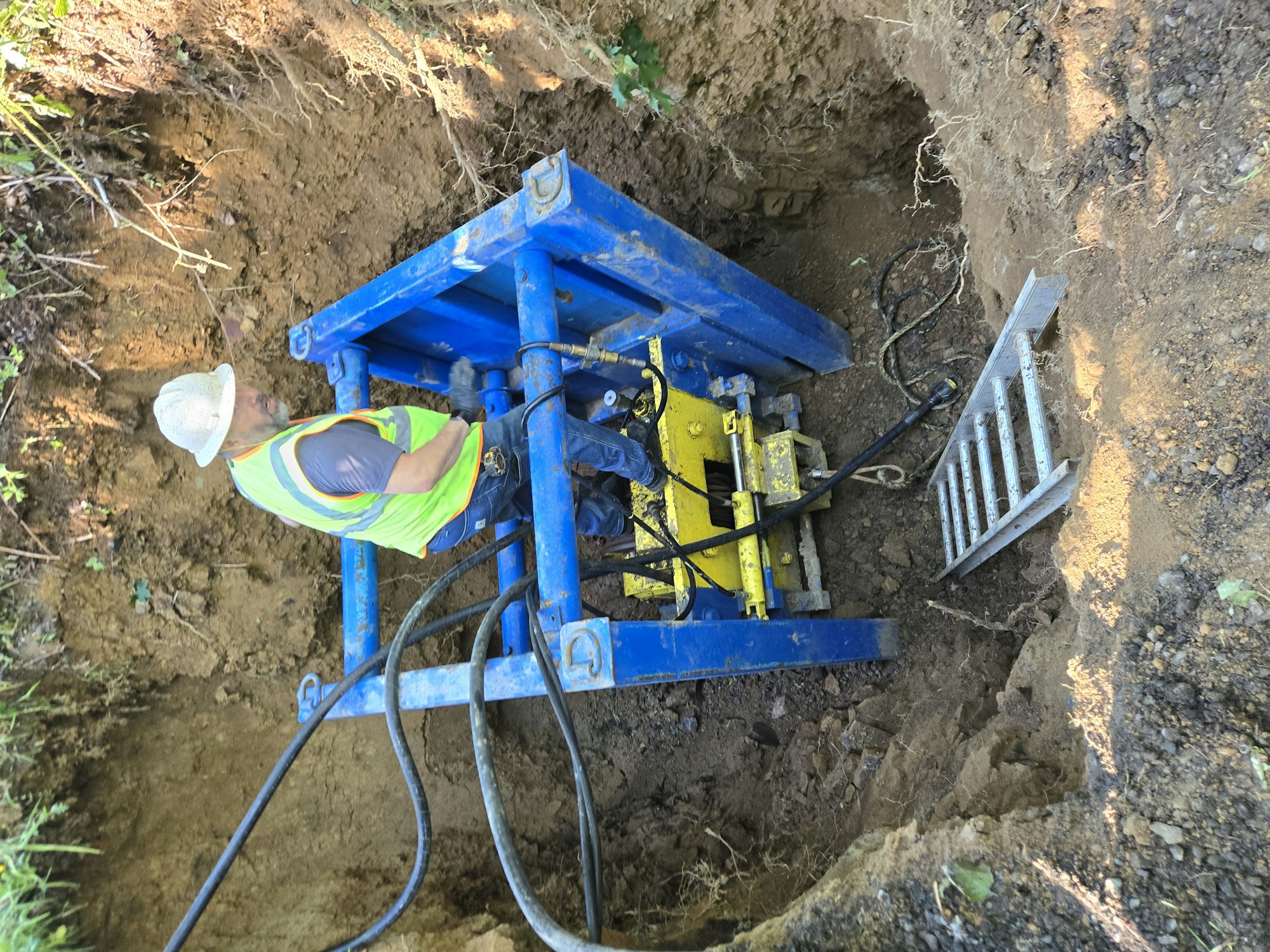 Construction worker operates machinery in a trench, wearing a safety vest and helmet. A metal ladder is nearby.