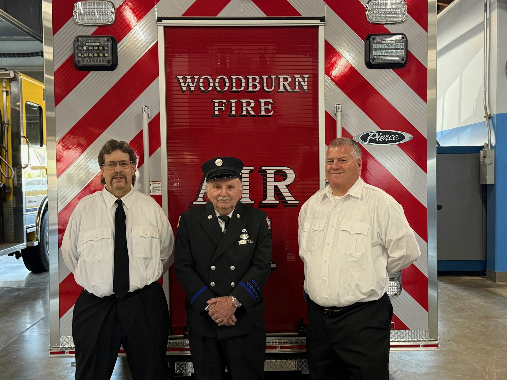 Three men, two in white shirts and one in a firefighter uniform, stand in front of a red and white Woodburn Fire ambulance.