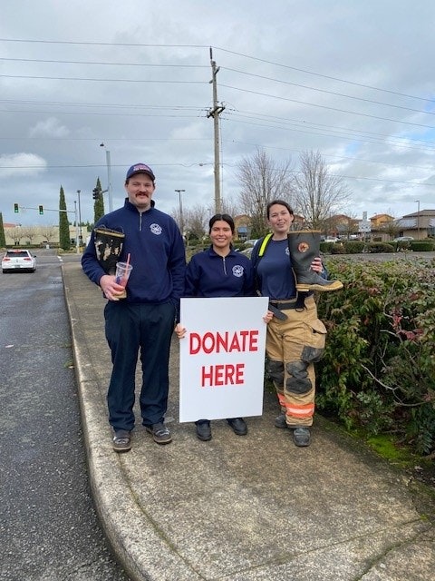 Three firefighters hold a "DONATE HERE" sign on the street, encouraging passersby to contribute.