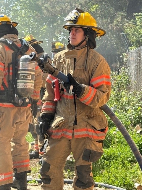 A firefighter in protective gear holds a hose, surrounded by fellow firefighters in a training or emergency response setting.