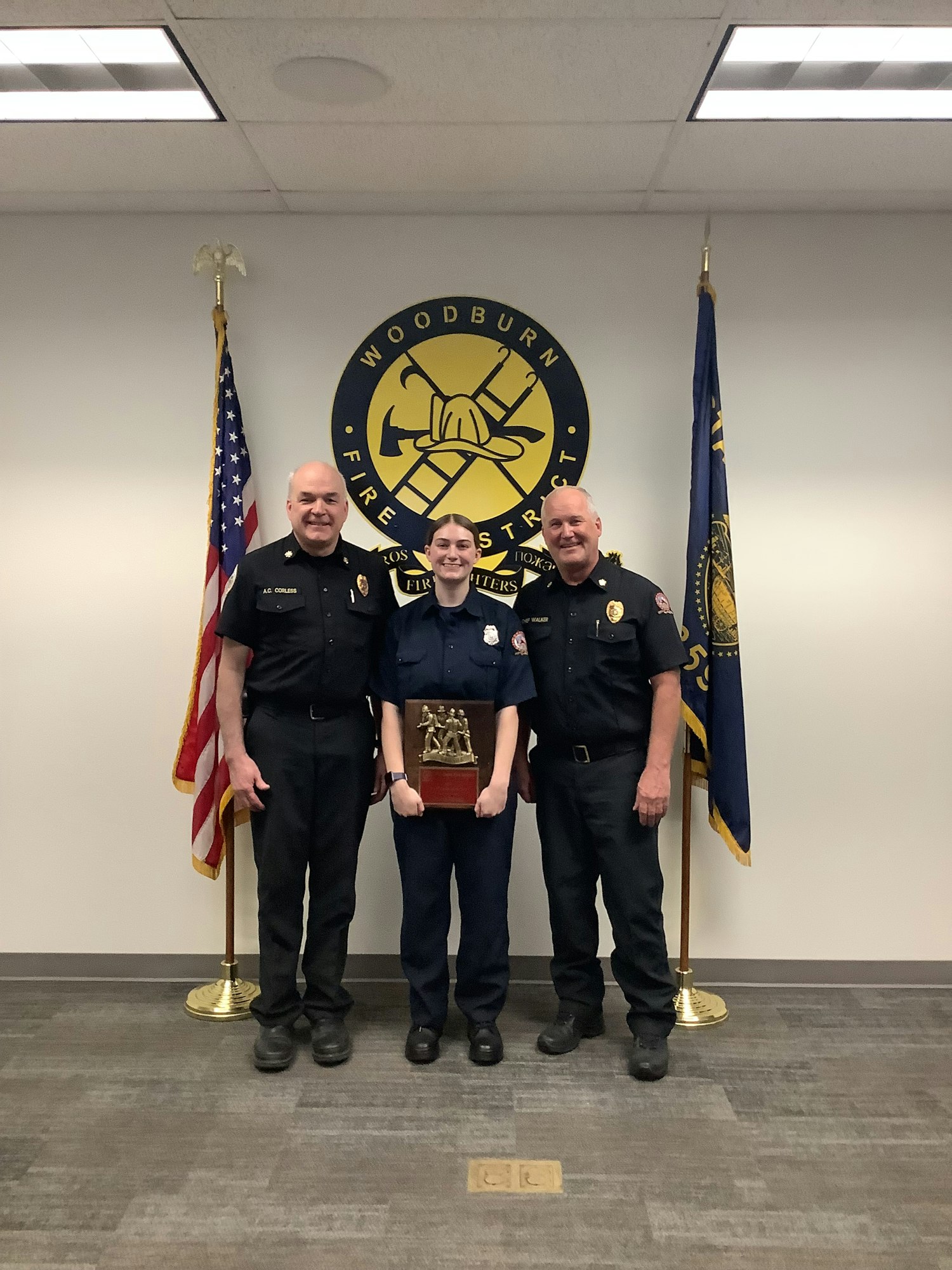 Three individuals in uniforms pose together in front of a fire station logo, with flags and a trophy in hand.