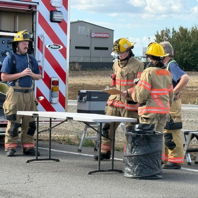 Firefighters in gear gather around a table near a fire truck, discussing or planning next steps in a response situation.