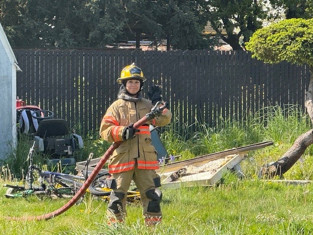 A firefighter stands in a grassy area, holding a hose, with equipment and a fence in the background.