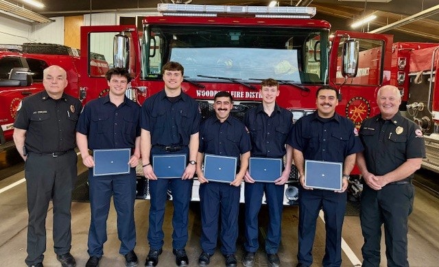 A group of firefighters stands proudly with certificates in front of a fire truck, celebrating their achievements.