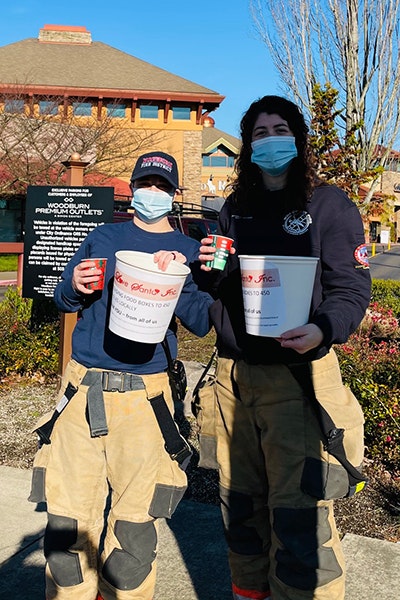 Two people in firefighter gear holding donation buckets and wearing masks, standing outdoors.