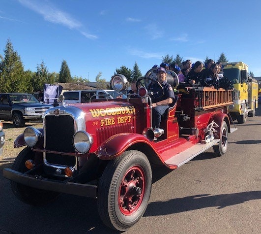A vintage red fire truck from Woodburn Fire Dept with people onboard, parked in a line of vehicles under a blue sky.