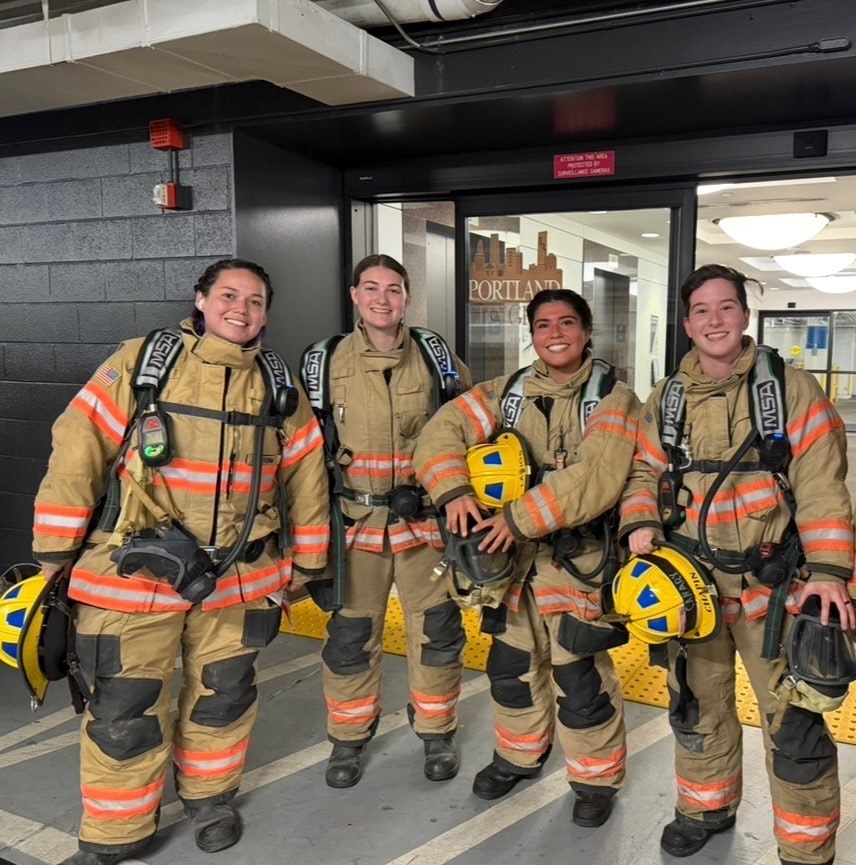 Four firefighters in gear, smiling and standing together in a hallway, showcasing camaraderie and readiness.