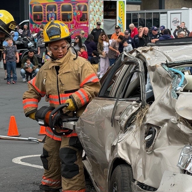 A firefighter uses tools to work on a severely damaged car while a crowd watches nearby at an event.