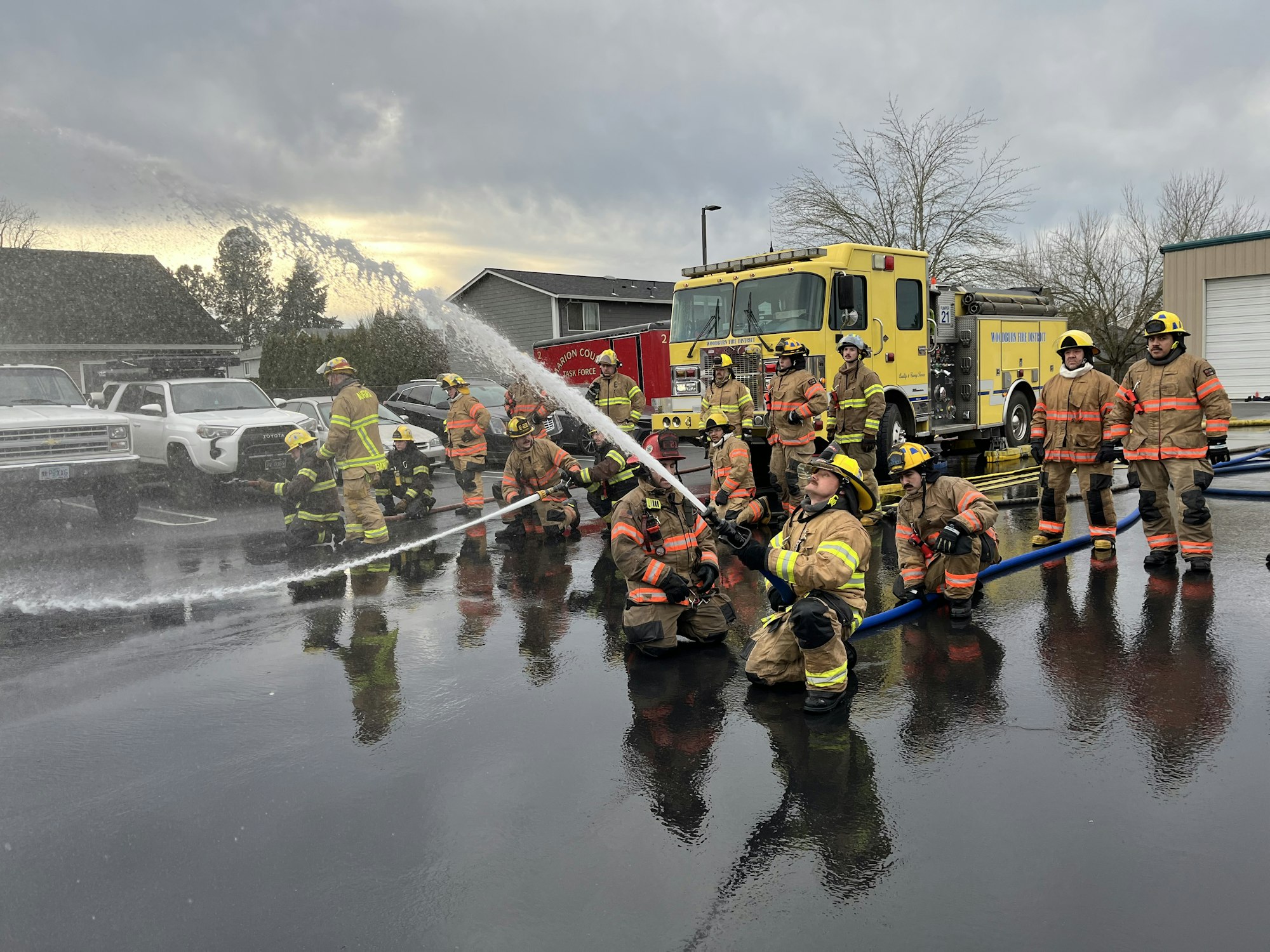 A group of firefighters training with hoses near their fire trucks, practicing water spraying techniques in a parking lot.