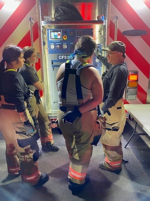 A group of four individuals in firefighting gear gather around a tool or equipment, discussing its use or operation.
